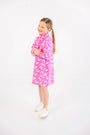 A young girl stands and smiles with her arms crossed, wearing The Beaufort Bonnet Company’s Tatum’s Turtleneck Dress in the Bows of Holly print—a bright pink, long-sleeve turtleneck dress made from soft Pima cotton featuring a white and green floral pattern—paired with white sneakers against a plain white background.