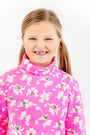 A smiling young girl with light brown hair in pigtails is wearing The Beaufort Bonnet Company’s Tatum’s Turtleneck Dress in the Bows of Holly print, standing in front of a white background.