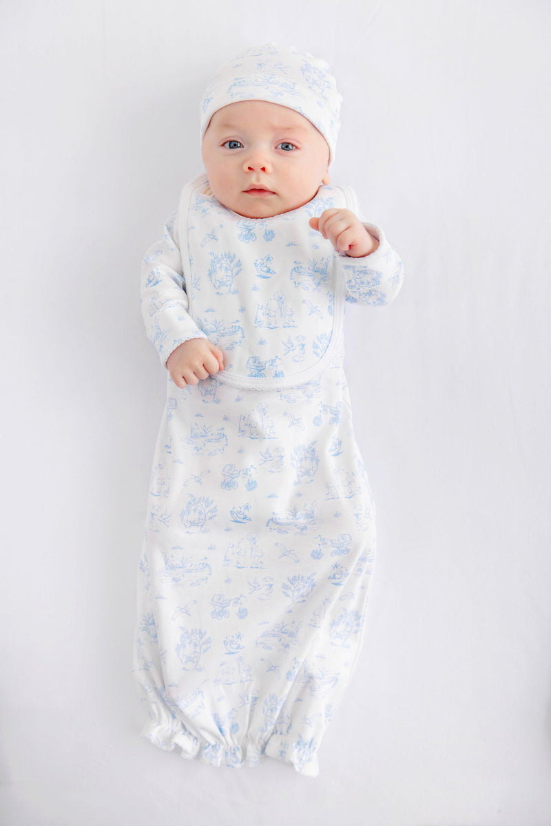 A baby dressed in The Beaufort Bonnet Company’s Sir Proper’s Sweet Arrival (Beale Street Blue) Bundle – a charming everyday set complete with a white hat, matching bib, and light blue layette gown – lies contentedly on a white background, gazing up with wide-eyed wonder. This adorable scene captures the perfect baby shower gift.