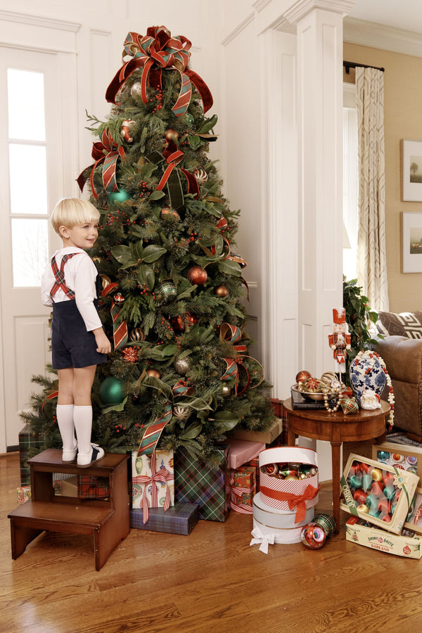 A young boy in Peabody Prep Short - Nantucket Navy Corduroy with Society Prep Plaid shorts stands on a step stool to decorate a Christmas tree.