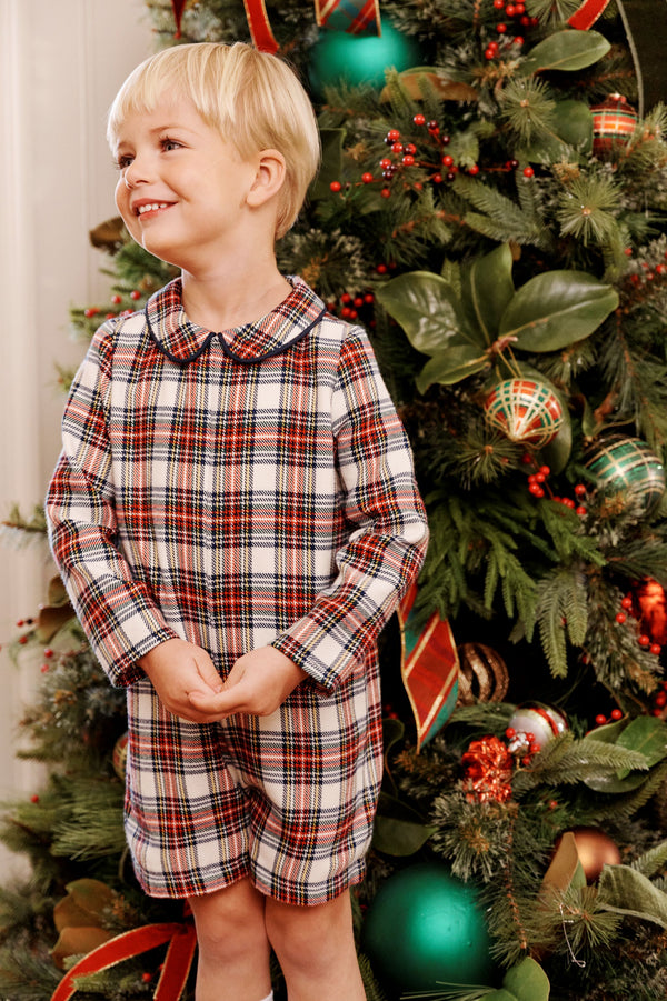 A young boy smiles in a John Hall Jon Jon - Paris Pike Plaid with Nantucket Navy long-sleeved romper with a rounded collar, posing in front of a decorated Christmas tree.