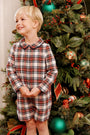 A young boy smiles in a John Hall Jon Jon - Paris Pike Plaid with Nantucket Navy long-sleeved romper with a rounded collar, posing in front of a decorated Christmas tree.