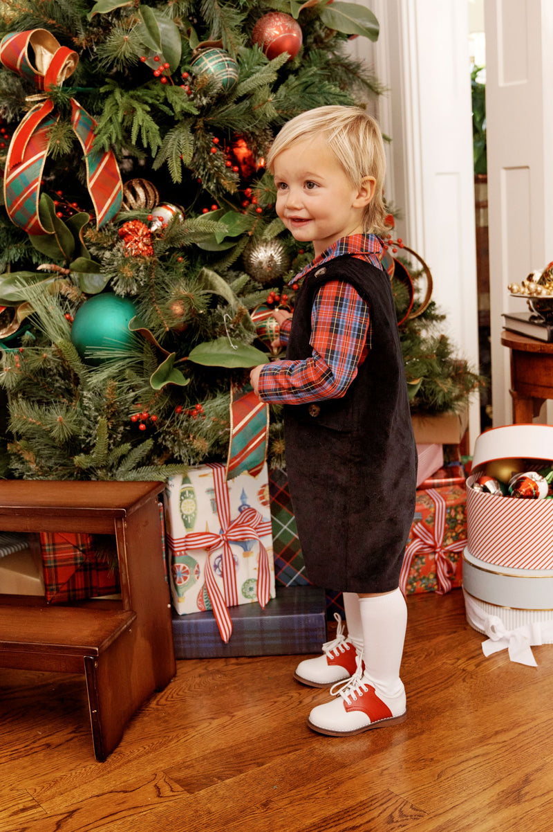 A young child dressed in The Beaufort Bonnet Company’s Jack Keene Jon Jon - Newport Night Corduroy stands by a decorated Christmas tree, smiling and holding an ornament. Wrapped presents and a box of ornaments are nearby on the wooden floor.