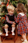 Two young children wearing The Beaufort Bonnet Company’s Long Sleeve Frenchy Frock in Bucktown Tartan with Worth Avenue White are by a decorated Christmas tree. One child sits on wooden steps, while the other stands nearby; both face away from the camera as festive ornaments and ribbons adorn the tree.