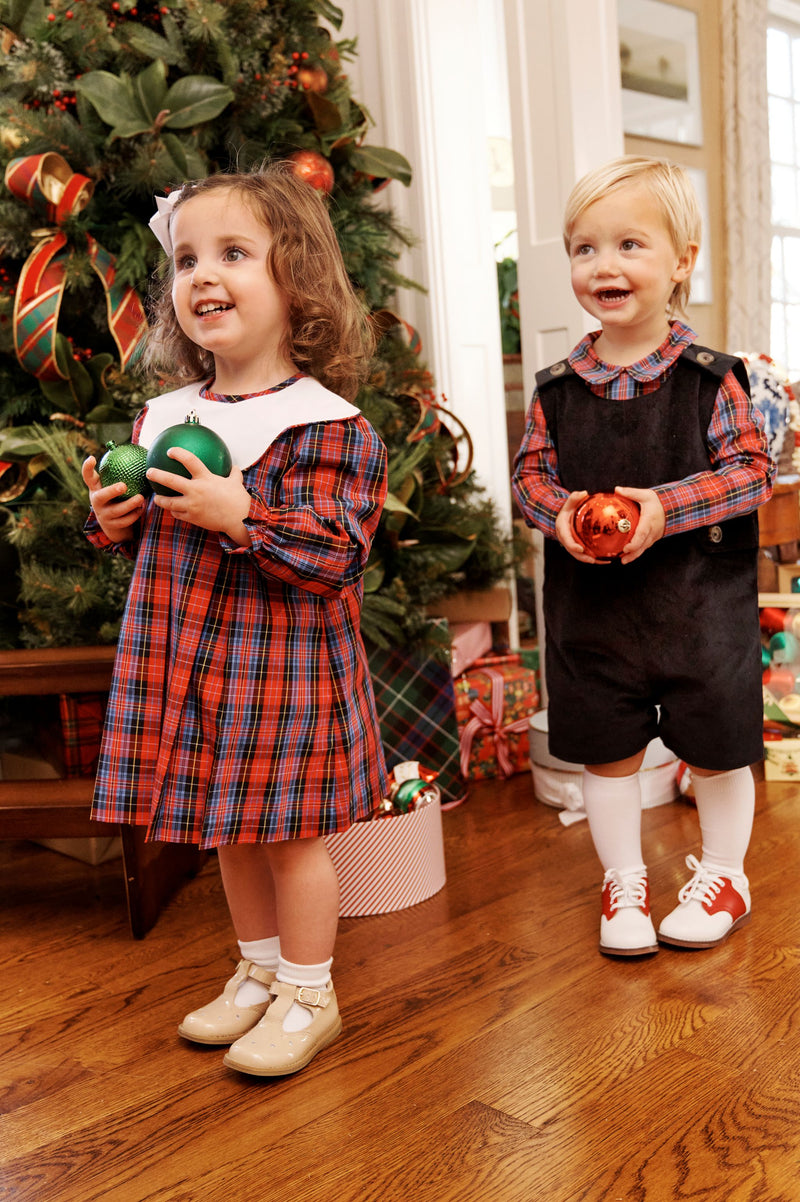 Two young children, dressed in the Long Sleeve Frenchy Frock - Bucktown Tartan with Worth Avenue White by The Beaufort Bonnet Company, stand smiling by a festively decorated Christmas tree. Each holds ornaments, and presents underneath the tree add to the joyful holiday atmosphere.