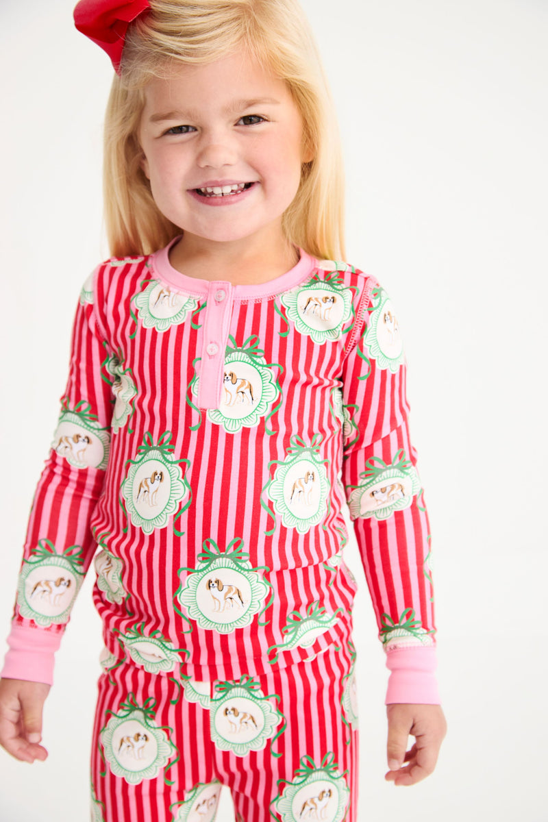 A young girl wears a Payton Pajama Set - Distinguished Doggies with Hamptons Hot Pink long-sleeved top and pants featuring a red and pink stripe pattern with dog illustrations inside decorative frames.