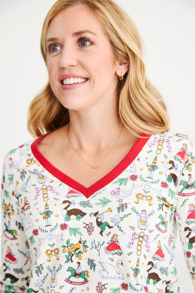 A smiling woman with blonde hair wears a white top from The Beaufort Bonnet Company’s Coastal Living Lounge Set – 12 Days of Christmas with Richmond Red, featuring colorful holiday-themed illustrations and red trim on the neckline, paired with a delicate gold necklace. The background is plain and light.