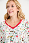 A smiling woman with blonde hair wears a white top from The Beaufort Bonnet Company’s Coastal Living Lounge Set – 12 Days of Christmas with Richmond Red, featuring colorful holiday-themed illustrations and red trim on the neckline, paired with a delicate gold necklace. The background is plain and light.