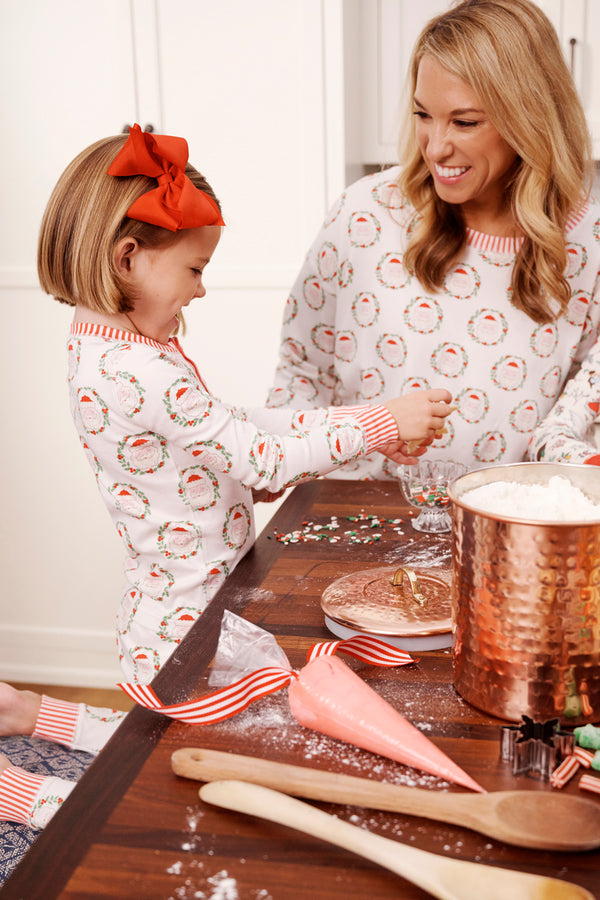 A smiling woman and young girl in matching Sutton’s Sweet Dream Set - North Pole Portrait with Richmond Red Stripe pajamas by The Beaufort Bonnet Company decorate cookies together at a kitchen table with frosting, sprinkles, and baking supplies.