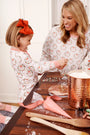 A smiling woman and young girl in matching Sutton’s Sweet Dream Set - North Pole Portrait with Richmond Red Stripe pajamas by The Beaufort Bonnet Company decorate cookies together at a kitchen table with frosting, sprinkles, and baking supplies.