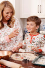 A woman and a boy, both dressed in The Beaufort Bonnet Company’s Sutton’s Sweet Dream Set - 12 Days of Christmas with Richmond Red pajamas, are smiling as they cut cookie dough with holiday-shaped cutters on the kitchen counter.