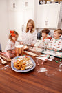 A woman and three children, all wearing The Beaufort Bonnet Company’s Snooze Button Set - North Pole Portrait with Richmond Red Stripe matching family pajamas, decorate cookies in a kitchen, surrounded by cookies, candy canes, and baking tools spread out on the wooden counter.