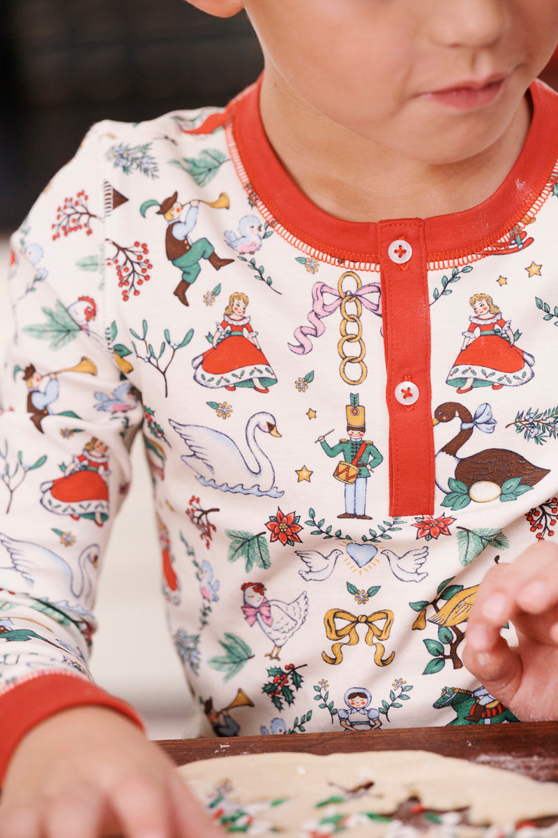 A child, dressed in The Beaufort Bonnet Company’s Sutton’s Sweet Dream Set - 12 Days of Christmas with Richmond Red—crafted from 100% Pima Cotton and adorned with festive illustrations of swans, nutcrackers, and dolls—is making crafts or cookies at a table.
