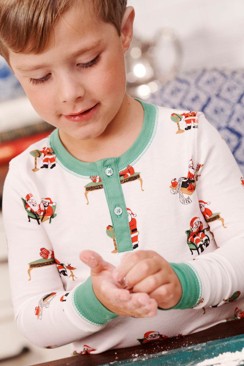 A young child in The Beaufort Bonnet Company’s Sutton’s Sweet Dream Set - Serious Santa Business kneads dough with both hands, focusing intently. The Pima cotton Christmas pajamas showcase playful Santa Claus designs, while the softly blurred background adds a gentle touch to the scene.