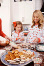 Wearing the Sutton’s Sweet Dream Set - Santa’s Laundry Line with Richmond Red pajamas from The Beaufort Bonnet Company, a woman and a young girl smile together as they decorate cookies in the kitchen, surrounded by baking tools, sprinkles, and an assortment of cookies spread out on the counter.