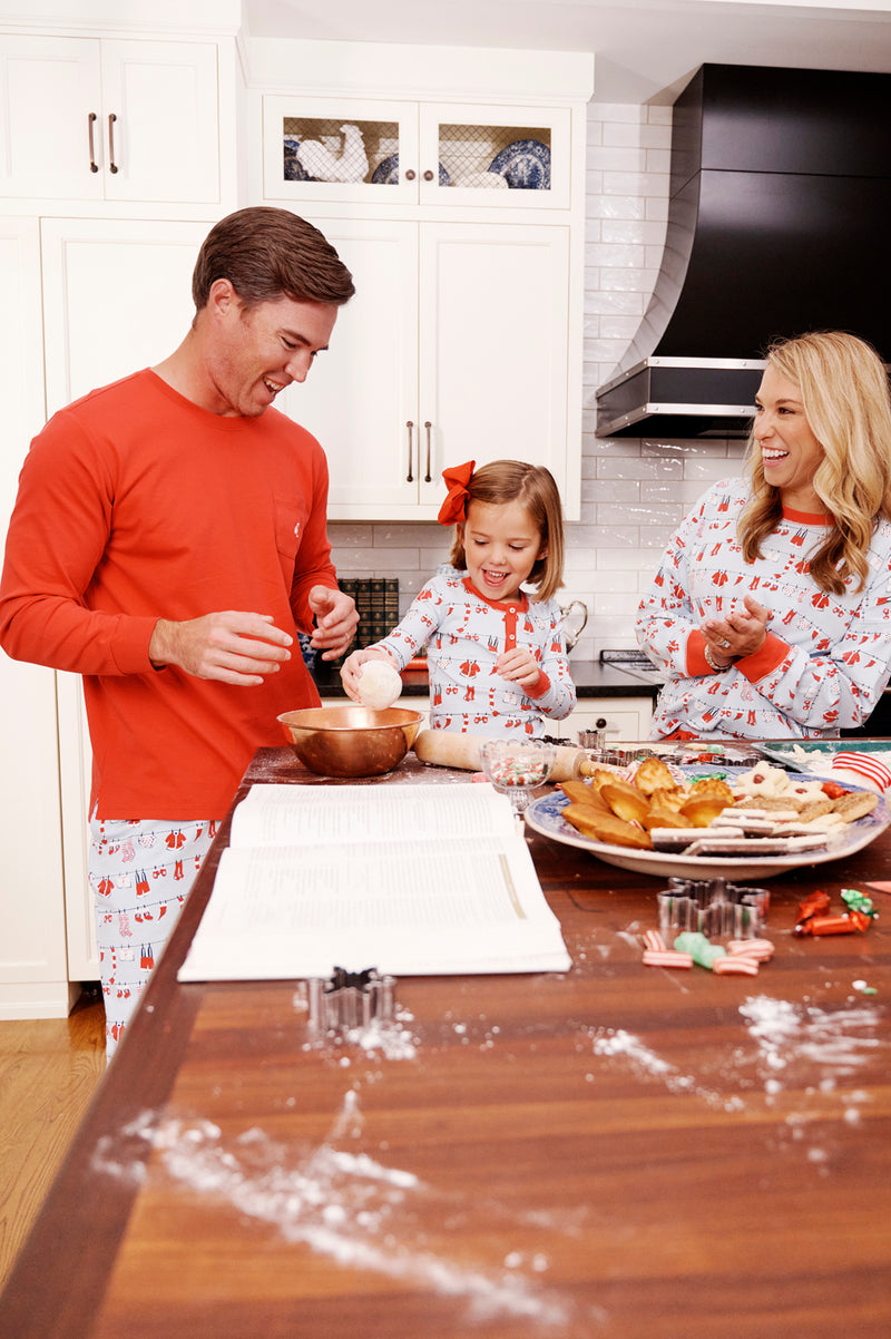 Wearing The Beaufort Bonnet Company’s Snooze Button Set - Santa’s Laundry Line, a family laughs together while baking cookies in a sunny kitchen. Dad mixes ingredients, mom beams nearby, and their daughter holds a ball of dough. Holiday treats and festive cookie cutters decorate the wooden counter.