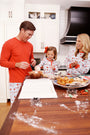 Wearing The Beaufort Bonnet Company’s Snooze Button Set - Santa’s Laundry Line, a family laughs together while baking cookies in a sunny kitchen. Dad mixes ingredients, mom beams nearby, and their daughter holds a ball of dough. Holiday treats and festive cookie cutters decorate the wooden counter.