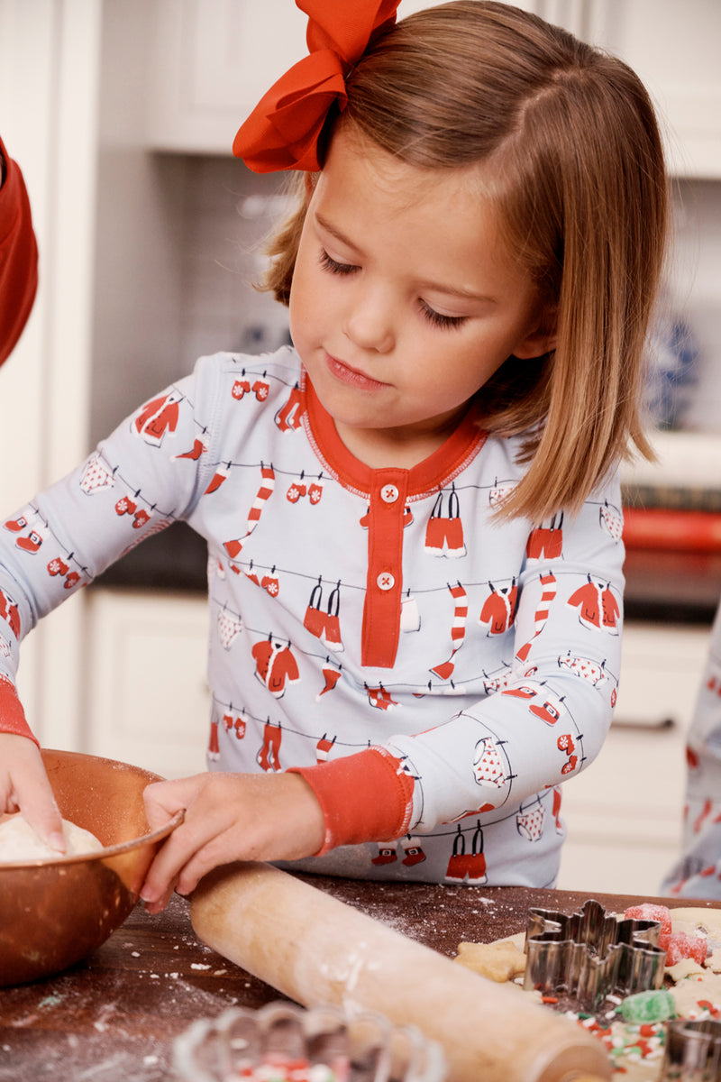 Wearing The Beaufort Bonnet Company’s Sutton’s Sweet Dream Set in the Santa’s Laundry Line with Richmond Red print, a young girl with a large red bow in her hair mixes dough in a bowl on a flour-dusted counter, surrounded by cookie cutters and a rolling pin as she prepares to bake cookies in the kitchen.