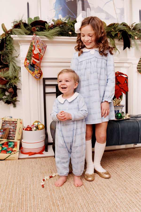 Two young children in matching blue pajamas stand before a festive fireplace adorned with garland, stockings, wrapped presents, and ornaments. The older girl beams in her The Beaufort Bonnet Company Long Sleeve Mary Dal Dress - Blue Choo Choos with Worth Avenue White, while the younger boy laughs, holding a red and white bead garland.