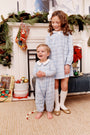 Two young children in matching blue pajamas stand before a festive fireplace adorned with garland, stockings, wrapped presents, and ornaments. The older girl beams in her The Beaufort Bonnet Company Long Sleeve Mary Dal Dress - Blue Choo Choos with Worth Avenue White, while the younger boy laughs, holding a red and white bead garland.