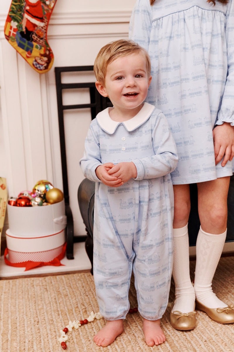 A young boy in a light blue Long Sleeve Bradford Romper - Blue Choo Choos with Worth Avenue White by The Beaufort Bonnet Company stands smiling next to an older girl in a matching dress, both posed in front of a fireplace adorned with Christmas stockings and ornaments.