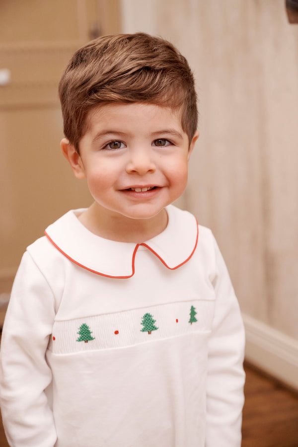 A smiling toddler wears the Samuel Smocked Romper - Worth Avenue White & Richmond Red with Trees, a long-sleeved white Pima cotton outfit featuring a peter pan collar with red trim and a chest panel embroidered with green trees and red dots.