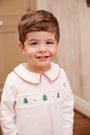 A smiling toddler wears the Samuel Smocked Romper - Worth Avenue White & Richmond Red with Trees, a long-sleeved white Pima cotton outfit featuring a peter pan collar with red trim and a chest panel embroidered with green trees and red dots.