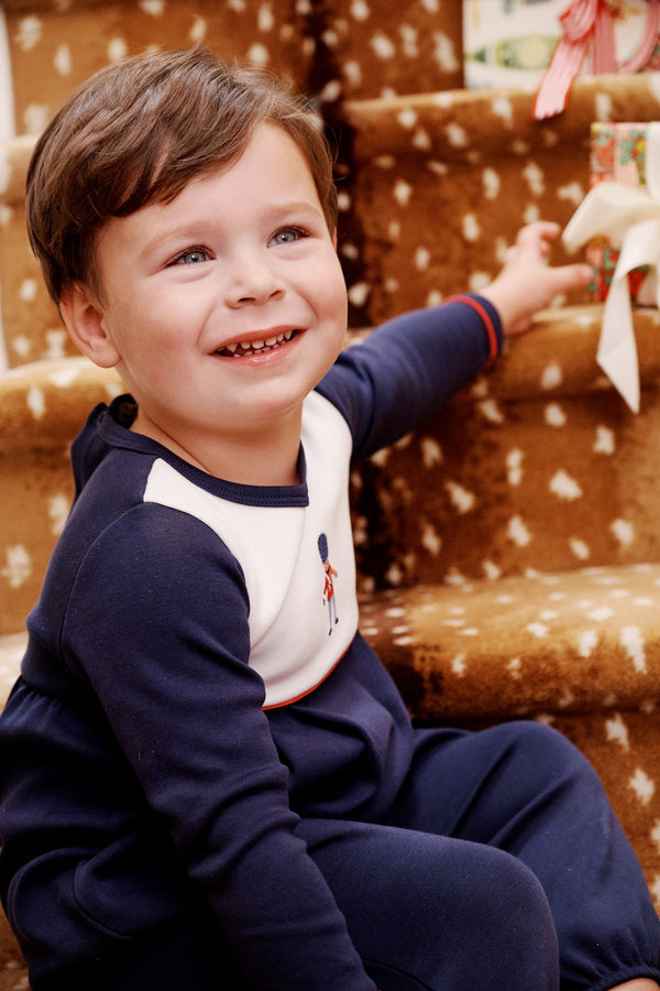 A young boy sits on stairs wearing a navy and white Long Sleeve Bradford Romper - Nantucket Navy & Worth Avenue White with Soldier Embroidery in Pima cotton knit with a small soldier embroidery on the chest.