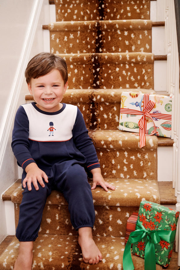 A smiling young boy wearing a The Beaufort Bonnet Company Long Sleeve Bradford Romper - Nantucket Navy & Worth Avenue White with Soldier Embroidery sits on a carpeted staircase next to two wrapped Christmas presents.
