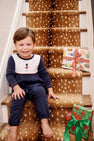 A smiling young boy wearing a The Beaufort Bonnet Company Long Sleeve Bradford Romper - Nantucket Navy & Worth Avenue White with Soldier Embroidery sits on a carpeted staircase next to two wrapped Christmas presents.