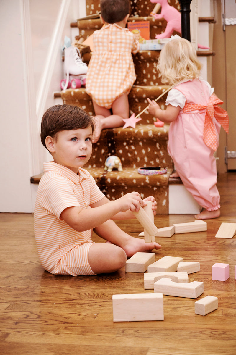 Three young children play indoors; one boy sits on the floor building with wooden blocks, while two girls wearing The Beaufort Bonnet Company’s Sir Proper’s Romper in Seashore Sherbet Stripe with Multicolor Stork enjoy playing with toys on and near carpeted stairs.
