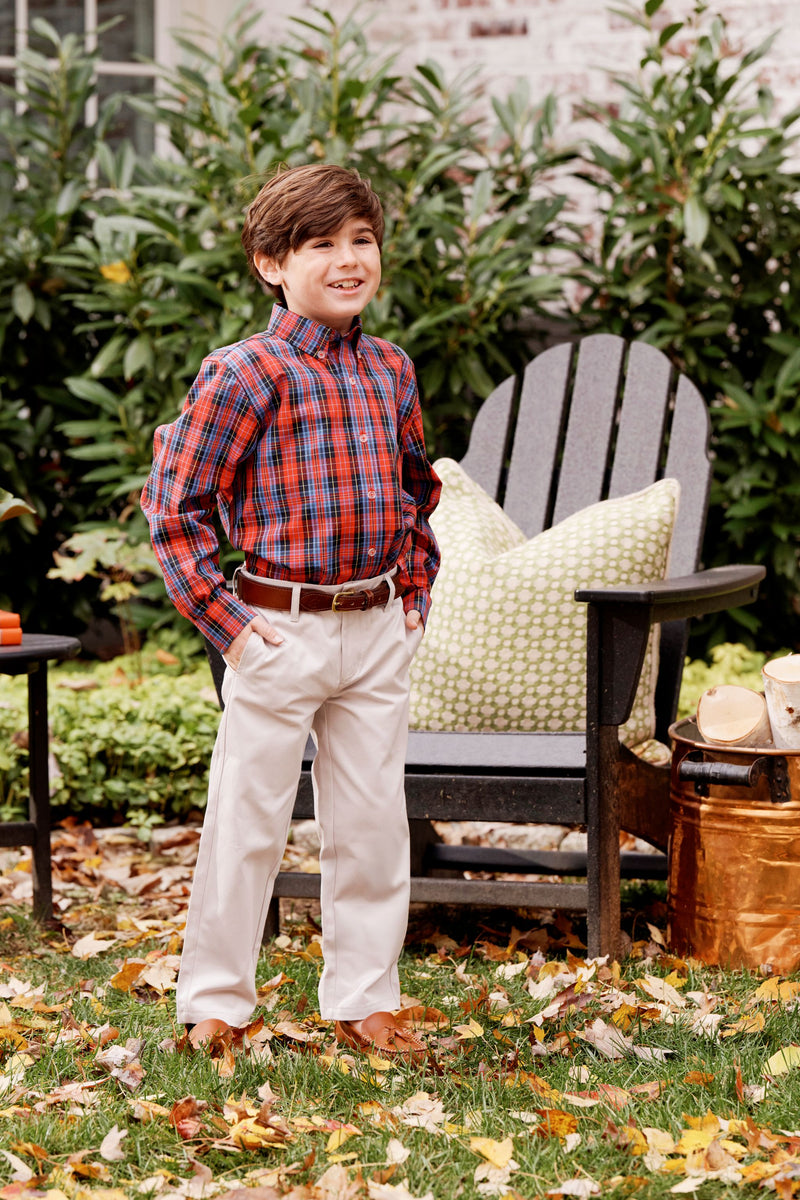 A young boy wearing The Beaufort Bonnet Company’s Dean’s List Dress Shirt in Bucktown Tartan with Richmond Red Stork and khaki pants stands smiling in a garden, next to an Adirondack chair featuring a green patterned pillow. Autumn leaves are scattered on the grass around him, creating a perfect setting for holiday gatherings.