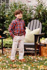 A young boy wearing The Beaufort Bonnet Company’s Dean’s List Dress Shirt in Bucktown Tartan with Richmond Red Stork and khaki pants stands smiling in a garden, next to an Adirondack chair featuring a green patterned pillow. Autumn leaves are scattered on the grass around him, creating a perfect setting for holiday gatherings.