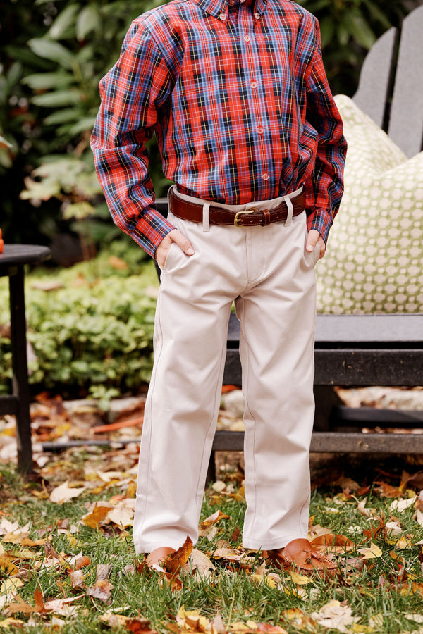 A child stands outdoors with hands in pockets, wearing a red plaid shirt, khaki pants accessorized with The Beaufort Bonnet Company’s Bullock Belt (Leather) in Tan, and brown shoes. Leaves cover the grass, and a chair with a green pillow is visible in the background.