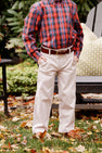 A child stands outdoors with hands in pockets, wearing a red plaid shirt, khaki pants accessorized with The Beaufort Bonnet Company’s Bullock Belt (Leather) in Tan, and brown shoes. Leaves cover the grass, and a chair with a green pillow is visible in the background.