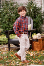 A smiling boy wearing a red plaid shirt and khaki pants accessorized with The Beaufort Bonnet Company’s Bullock Belt (Leather) in Tan stands outside with his hands in his pockets, surrounded by fallen leaves. A chair and firewood can be seen in the background.