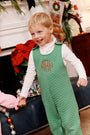 A smiling young boy wears The Beaufort Bonnet Company’s Tatum’s Turtleneck & Onesie (Unisex) in Worth Avenue White, holding hands with someone just out of frame. Festive holiday decorations, including a poinsettia and a Christmas stocking, decorate the background.