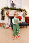 A young child in a holiday outfit—a Lawson Longall in Kiawah Kelly Green Quilted by The Beaufort Bonnet Company and red shoes—smiles while holding a popcorn and cranberry garland in a festive living room with holiday decorations, stockings, and toys near a decorated fireplace mantel.