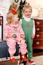 Two young children in festive holiday outfits smile indoors. The girl wears The Beaufort Bonnet Company’s Long Sleeve Ramona Ruffle Romper in Hamptons Hot Pink with Wreath Embroidery and holds a garland, accented by a red bow; the boy is dressed in a green quilted outfit with a monogram. Holiday decorations are visible in the background.