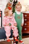 Two young children in festive holiday outfits smile indoors. The girl wears The Beaufort Bonnet Company’s Long Sleeve Ramona Ruffle Romper in Hamptons Hot Pink with Wreath Embroidery and holds a garland, accented by a red bow; the boy is dressed in a green quilted outfit with a monogram. Holiday decorations are visible in the background.