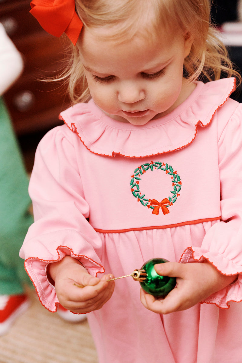 A young child wearing The Beaufort Bonnet Company’s Long Sleeve Ramona Ruffle Romper in Hamptons Hot Pink with Wreath Embroidery holds a green Christmas ornament, looking down at it.