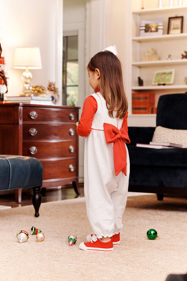 A young girl dressed in The Beaufort Bonnet Company’s Sleeveless Rebecca Romper—featuring Kiawah Kelly Green Microdot with Richmond Red—stands in a living room, facing away, while Christmas ornaments are scattered on the floor around her.