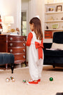 A young girl dressed in The Beaufort Bonnet Company’s Sleeveless Rebecca Romper—featuring Kiawah Kelly Green Microdot with Richmond Red—stands in a living room, facing away, while Christmas ornaments are scattered on the floor around her.