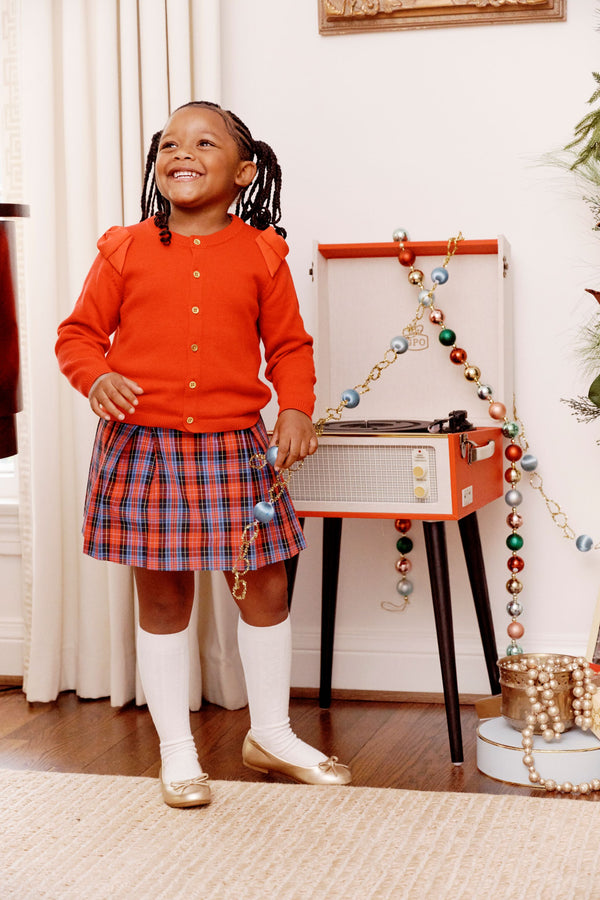 A smiling young girl wears The Beaufort Bonnet Company’s Cambridge Cardigan in Richmond Red with bows, paired with a plaid skirt, as she stands by a record player decorated with colorful beads and holds a string of beads. She completes her look with white knee socks and gold shoes—a charming addition to any little one’s wardrobe.