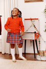 A smiling young girl wears The Beaufort Bonnet Company’s Cambridge Cardigan in Richmond Red with bows, paired with a plaid skirt, as she stands by a record player decorated with colorful beads and holds a string of beads. She completes her look with white knee socks and gold shoes—a charming addition to any little one’s wardrobe.