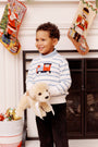 A young child smiles while holding a plush dog. He wears The Beaufort Bonnet Company’s “Isaac’s Intarsia Sweater - Barrington Blue Stripe with Train.” Behind him are Christmas stockings, ornaments in a white container, and a fireplace.