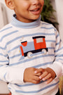 A young child smiles, wearing The Beaufort Bonnet Company’s Isaac’s Intarsia Sweater in Barrington Blue Stripe with a colorful train motif on the front. The child’s hands are held together in front of their body.