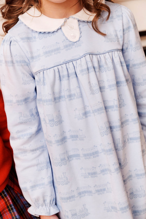 A child wearing The Beaufort Bonnet Company’s Long Sleeve Mary Dal Dress in Blue Choo Choos with Worth Avenue White—a light blue, long-sleeve dress featuring a white collar and a playful train pattern—stands next to another person partly visible in red.