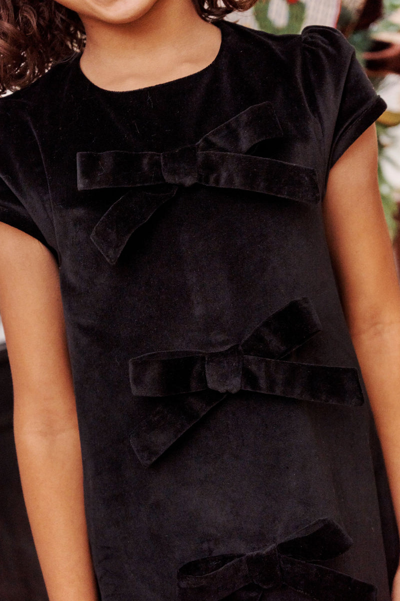 A child wearing The Beaufort Bonnet Company’s Betts Bow Dress - Newport Night Velveteen, a short-sleeved velveteen dress featuring three large velvet bows on the front. Only the lower face, shoulders, and upper body are visible.