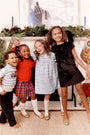 Four smiling children stand together in front of a decorated fireplace with stockings and garland, dressed in festive clothing including The Beaufort Bonnet Company’s Cambridge Cardigan in Richmond Red with Bows, posing for a holiday photo that’s perfect for adding to your little one’s wardrobe memories.